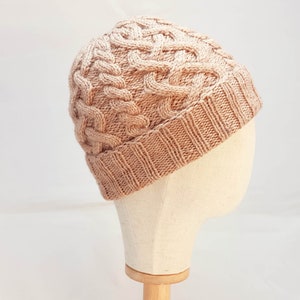 May include: A light brown cable-knit beanie hat. The hat features a textured, woven design with a ribbed brim. The hat is displayed on a mannequin head against a white background. The hat is a warm, cozy accessory.