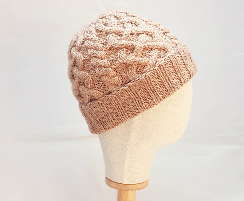 May include: A light brown cable-knit beanie hat. The hat features a textured, woven design with a ribbed brim. The hat is displayed on a mannequin head against a white background. The hat is a warm, cozy accessory.