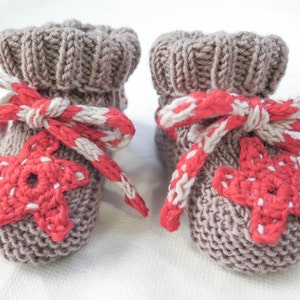 Puede incluir: Un par de patucos de bebé de punto en color marrón suave. Cada patuco presenta una aplicación de estrella roja y un lazo a rayas rojas y blancas. Los patucos están diseñados con un puño acanalado.