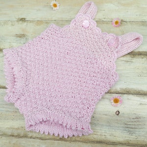 May include: A pink knitted baby romper with a scalloped neckline and two buttons. The romper is lying on a light wood surface.