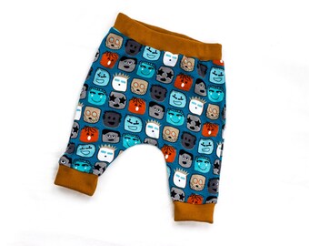 Op de afbeelding: Blauwe en oranje babybroek met een patroon van kleurrijke gezichten. De broek heeft een bruine elastische tailleband.
