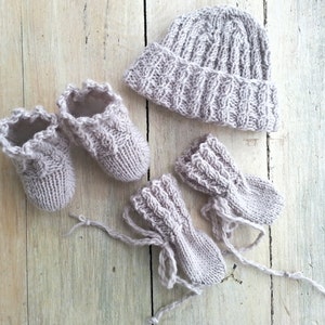 May include: A set of knitted baby clothes in a light beige color. The set includes a hat, booties, and mittens.