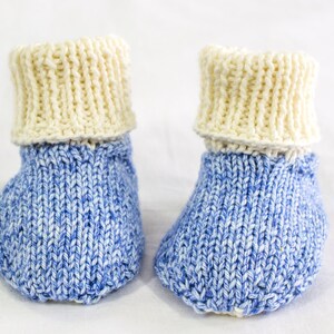 May include: A pair of hand-knitted baby booties. The booties have a blue and white speckled pattern on the foot and a cream-colored cuff. The soft, textured knitwear is designed for infants.