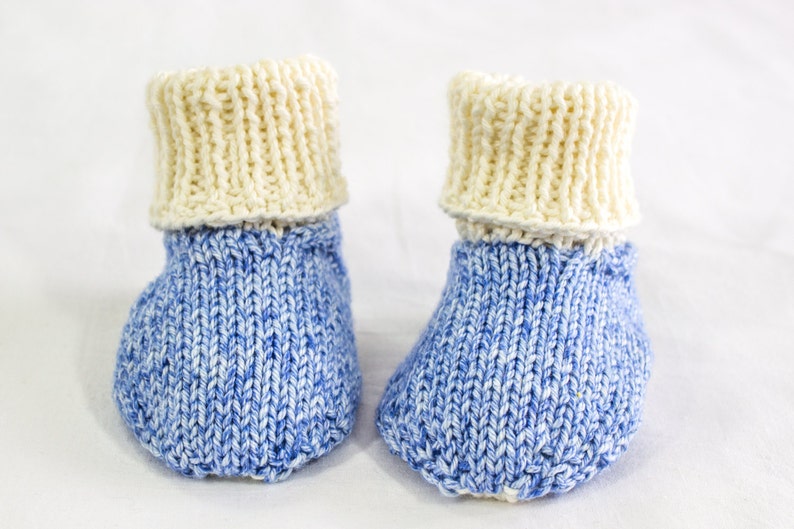 May include: A pair of hand-knitted baby booties. The booties have a blue and white speckled pattern on the foot and a cream-colored cuff. The soft, textured knitwear is designed for infants.