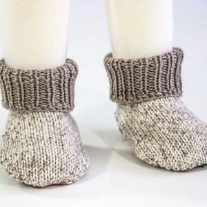 May include: A pair of knitted baby booties. The booties are primarily white with flecks of gray, and have a ribbed, dark gray cuff. The booties are designed to keep a baby's feet warm and cozy.