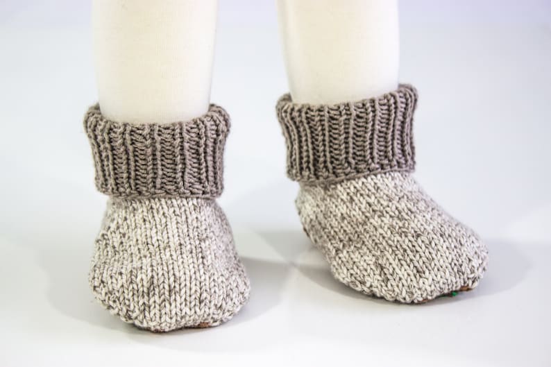 May include: A pair of knitted baby booties. The booties are primarily white with flecks of gray, and have a ribbed, dark gray cuff. The booties are designed to keep a baby's feet warm and cozy.