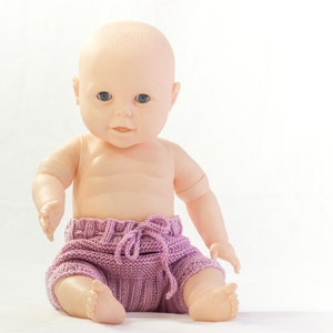 Puede incluir: Una muñeca de bebé de plástico con un pañal de punto morado. La muñeca tiene ojos azules y una tez rosa claro.