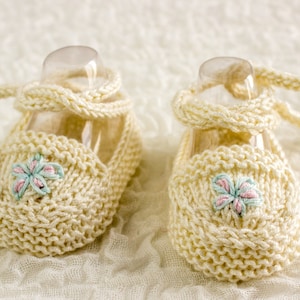 May include: A pair of knitted baby booties with a light pink and blue flower embroidery on each bootie. The booties are cream colored and have a lace-up design.
