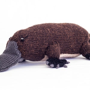 Puede incluir: Un juguete de peluche de ornitorrinco tejido de color marrón y gris. El ornitorrinco tiene un pico largo y plano y patas palmeadas.
