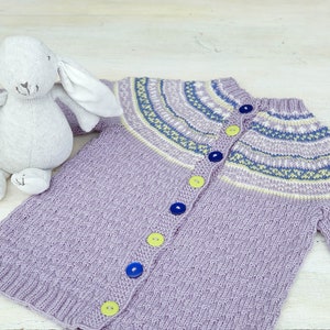 May include: A purple knitted baby cardigan with a colourful patterned yoke and button closure. The cardigan has yellow and blue buttons.