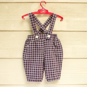 May include: Red plaid overalls with a button closure. The overalls are hanging on a red plastic hanger.