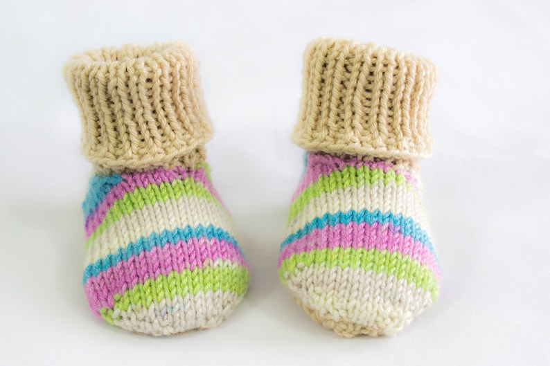 May include: A pair of hand-knitted baby booties. The cuffs are a solid beige color, while the foot of the booties features stripes of white, pink, blue, and green. The booties are made of soft yarn and are designed for comfort.