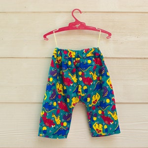 May include: A pair of teal blue children's pants with a colorful dinosaur print. The pants have an elastic waistband and a drawstring.