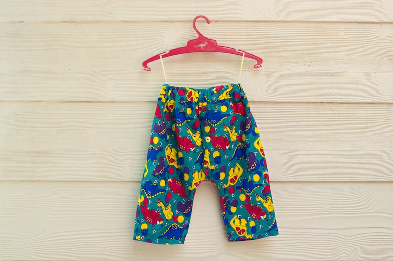 May include: A pair of teal blue children's pants with a colorful dinosaur print. The pants have an elastic waistband and a drawstring.