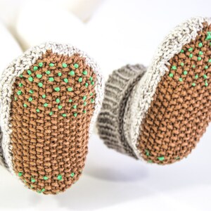May include: A pair of knitted baby booties in brown, gray, and white. The soles are brown with small green dots for grip. The cuffs are gray and white, and the booties are shown against a white background.