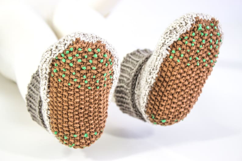 May include: A pair of knitted baby booties in brown, gray, and white. The soles are brown with small green dots for grip. The cuffs are gray and white, and the booties are shown against a white background.