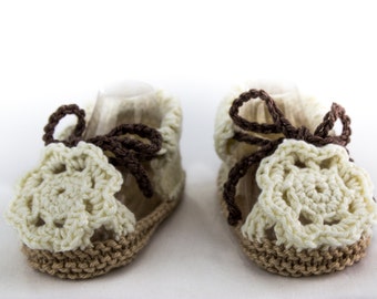 May include: A pair of crocheted baby sandals in cream and brown. The sandals have a flower design and a brown ribbon.