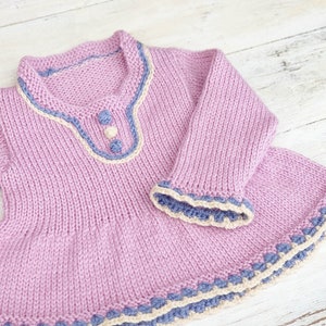 May include: A hand-knitted, light purple baby dress with long sleeves. The dress features a round neckline and cuffs trimmed with blue and cream-colored yarn. Decorative buttons adorn the bodice, and the hemline is also trimmed with blue and cream yarn.