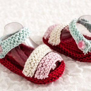 Puede incluir: Un par de sandalias de bebé de punto en rojo, blanco, rosa y azul con un detalle de botón.