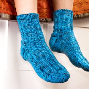 Puede incluir: Un par de calcetines azules de punto con un patr&oacute;n texturizado. Los calcetines est&aacute;n puestos en los pies de una persona y cuelgan del borde de una superficie blanca.