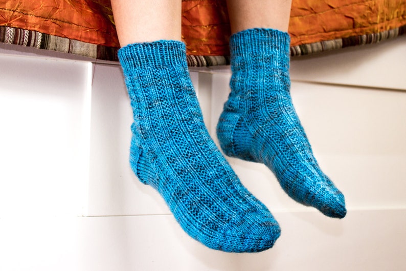 Puede incluir: Un par de calcetines azules de punto con un patr&oacute;n texturizado. Los calcetines est&aacute;n puestos en los pies de una persona y cuelgan del borde de una superficie blanca.