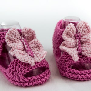 May include: A pair of pink knitted baby sandals with butterfly embellishments. The sandals have a strap with a button closure and an open toe design.