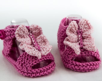 May include: A pair of pink knitted baby sandals with butterfly embellishments. The sandals have a strap with a button closure and an open toe design.