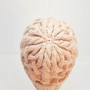 May include: A light brown knitted beanie hat with a cable knit design. The hat is made of a soft, textured yarn and has a round crown. The hat is a neutral color and would be suitable for fall or winter wear.
