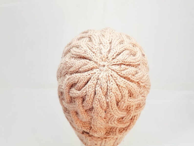 May include: A light brown knitted beanie hat with a cable knit design. The hat is made of a soft, textured yarn and has a round crown. The hat is a neutral color and would be suitable for fall or winter wear.