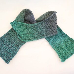 May include: A green and gray knitted scarf with a gradient color pattern. The scarf is folded in half and tied in a knot.