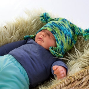 Peut inclure: Un bébé portant un bonnet tricoté bleu et vert avec des pompons, une chemise bleue et un pantalon bleu et vert, dormant dans un panier en osier doublé de fausse fourrure blanche.