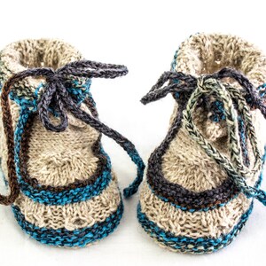 Puede incluir: Un par de botitas de bebé tejidas con base beige y detalles en azul, marrón y verde. Las botitas tienen un cierre de cordón.