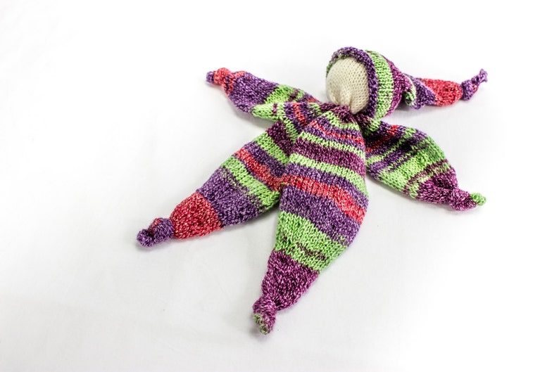 May include: A handmade knitted doll with a striped purple, green, and red outfit. The doll has a white fabric head and a knitted hat.