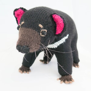 May include: A black and brown knitted toy Tasmanian devil with pink ears and white markings on its chest. The toy is standing on a white surface.
