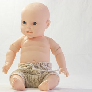 Puede incluir: Una muñeca de plástico con ojos azules, que lleva un pañal de punto beige. La muñeca está sentada sobre una superficie blanca.