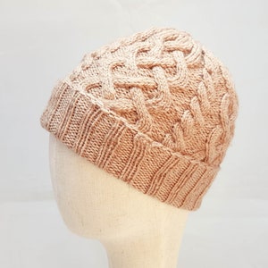May include: A light brown knitted beanie hat with a cable knit design. The hat is made of a soft, textured yarn and has a folded brim. The hat is displayed on a mannequin head against a white background.