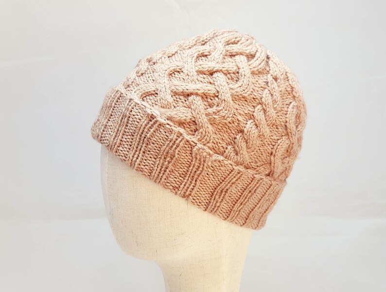 May include: A light brown knitted beanie hat with a cable knit design. The hat is made of a soft, textured yarn and has a folded brim. The hat is displayed on a mannequin head against a white background.
