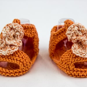 May include: A pair of orange knitted baby sandals with white and orange crocheted flower accents.