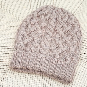 May include: A mauve-coloured knitted beanie hat. The hat has a cable knit pattern on the crown and a ribbed brim. The hat is made from a soft, textured yarn and is displayed on a woven surface.