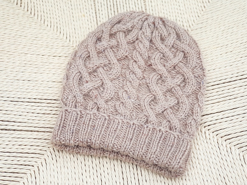 May include: A knitted beanie in a light mauve color. The hat features a cable knit design on the crown and a ribbed brim. The hat is made of a soft, textured yarn and is displayed on a woven surface.