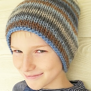 Puede incluir: Un gorro de punto a rayas azules, marrones y beige con un borde doblado. El gorro lo lleva puesto un niño con el pelo rubio.