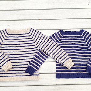 Könnte beinhalten: Zwei gestreifte Pullover, einer in Beige und Marineblau, der andere in Marineblau und Beige, auf einer weißen Holzoberfläche ausgelegt.