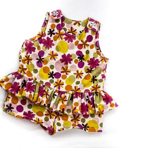 May include: A white sleeveless romper with a ruffled hem. The romper has a floral print with pink, yellow, and green flowers and circles.