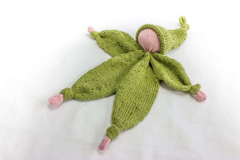 May include: A green knitted doll with pink accents. The doll is lying on its back with its arms and legs outstretched. The doll has a knitted hat and a knitted face.