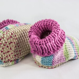 May include: A pair of handmade knitted baby booties. The booties feature a beige base with pink, blue, and green accents. The cuffs are a vibrant pink. The booties are designed for comfort and warmth.