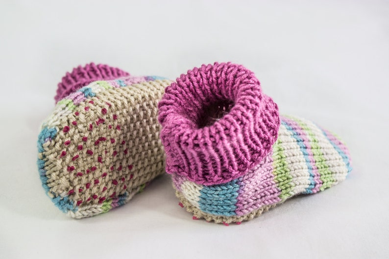 May include: A pair of handmade knitted baby booties. The booties feature a beige base with pink, blue, and green accents. The cuffs are a vibrant pink. The booties are designed for comfort and warmth.