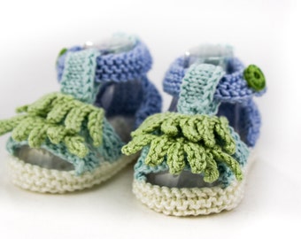 May include: A pair of knitted baby sandals in blue, green, and white. The sandals have a fringe detail and a button on the strap.