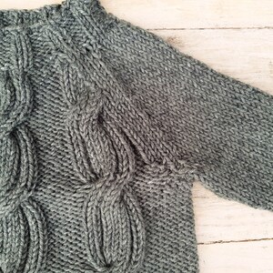May include: A gray knitted sweater with a cable knit pattern. The sweater has a crew neck and long sleeves.