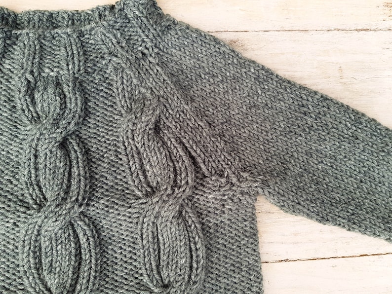 May include: A gray knitted sweater with a cable knit pattern. The sweater has a crew neck and long sleeves.
