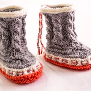 May include: A pair of gray knitted baby booties with white and orange trim. The booties have a cable knit pattern and are tied with orange and white yarn.
